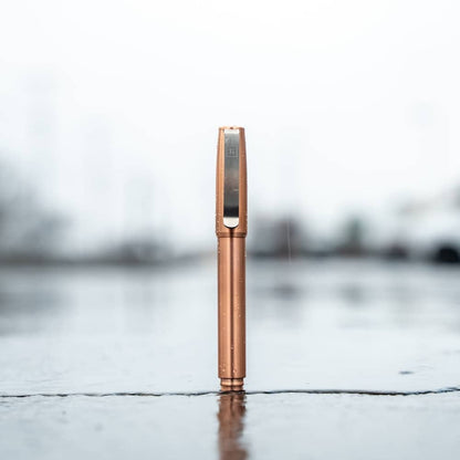 Copper titanium capped pen by BIG IDEA DESIGN LLC standing upright on wet surface with blurred background